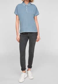 Vaaleansininen puuvillapolo, jossa on kaulus ja kolme nappia, yhdistettynä mustiin kapeisiin farkkuihin ja valkoisiin lenkkareihin.