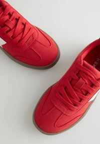 Czerwone trampki wykonane z zamszowego materiału, z okrągłymi noskami, płaskimi brązowymi gumowymi podeszwami i pasującymi sznurowadłami z białym akcentem po boku.