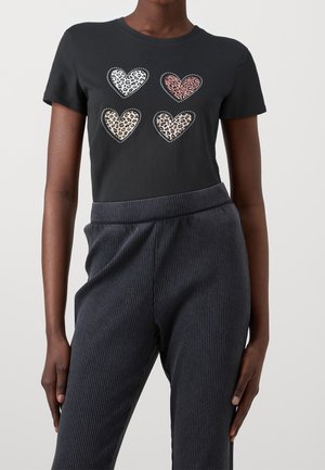 Chemise noire à manches courtes avec quatre cœurs imprimés léopard en blanc, rose, beige et marron, portée avec un pantalon côtelé foncé.