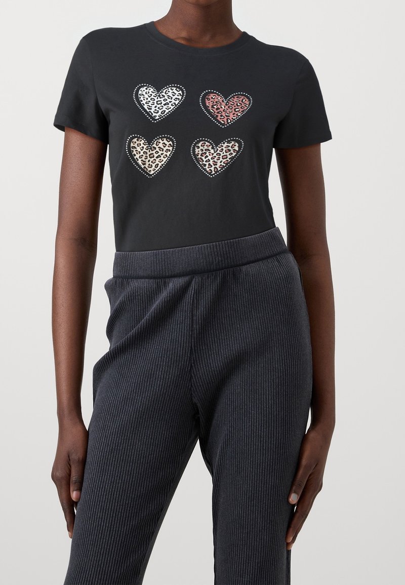 Chemise noire à manches courtes avec quatre cœurs imprimés léopard en blanc, rose, beige et marron, portée avec un pantalon côtelé foncé.