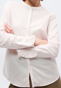 Femme portant un chemisier rose pâle à manches longues avec des poignets volantés et un col boutonné, les bras croisés sur la poitrine.