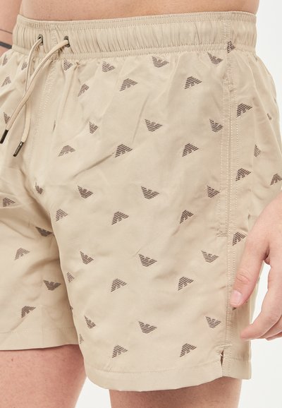 Shorts da bagno beige con un motivo sottile di piccole forme triangolari marroni. Presentano una vita elastica con cordoncino e dettagli con fessure laterali.