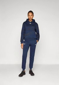 Tmavomodrá mikina s kapucňou, klokanie vrecko, šnúrky a jemné vzorované detaily, doplnená o zodpovedajúce nohavice so značkovým logom. Čierne tenisky.