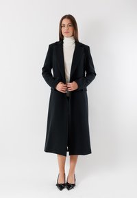 Cappotto lungo nero con rever a punta e doppi bottoni, realizzato in un tessuto liscio, abbinato a un dolcevita bianco e a scarpe con tacco nere.