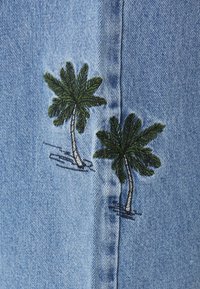 Hele sinine denim kangas, millel on kaks rohelise ja hõbedase värviga tikandiga palmipuud, detailsete lehtede ja lühikeste tüvedega.
