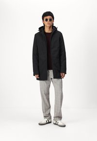 Μαύρο αδιάβροχο jacket με κουκούλα, μπορντό πουκάμισο, καρό γκρι παντελόνι και λευκά αθλητικά παπούτσια με μαύρες ρίγες. Προστέθηκαν στρογγυλά γυαλιά ηλίου.