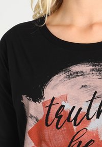 Schwarzes Baumwoll-T-Shirt mit einem grafischen Druck, der abstrakte rosa und rote Pinselstriche sowie das Wort "Wahrheit" in fettem schwarzen Text zeigt.