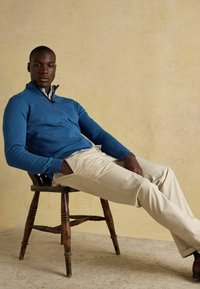 Zils adījuma džemperis ar rāvējslēdzēja kaklu, kopā ar gaiši bēšām chinos biksēm. Vīrietis ērtā pozā sēž uz koka krēsla neitrālas krāsas fonā.