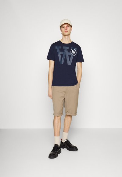 Tricou din bumbac bleumarin cu logo-uri mari gri, pantaloni scurți bej cu manșete rulante, șosete crem și pantofi negri cu șireturi și talpă texturată.