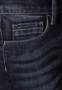 Jeans de mezclilla azul oscuro con un pequeño bolsillo trasero y detalles de costura amarilla. La tela tiene una textura suave y patrones de sombreado sutiles.