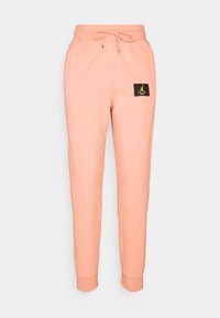 Peach joggers made from soft fabric, featuring a drawstring waist, side pockets, and a black logo patch with yellow accents.