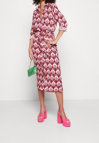 Czerwona i różowa sukienka w wzory z talią stałą, rękawami o długości trzech kwart, i kołnierzykiem, zestawiona z różowymi platformami i zieloną torebką.