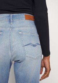 Ljusblå denimjeans med en brun läderlapp på bakre midjan, två bakfickor och en zigzag-sticksöm.