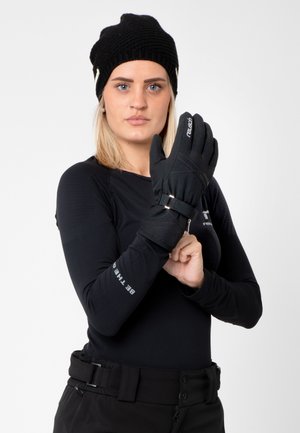 Black snow gloves with textured fabric, adjustable wrist strap, and brand logo, worn by a person in a black long-sleeve shirt and hat.