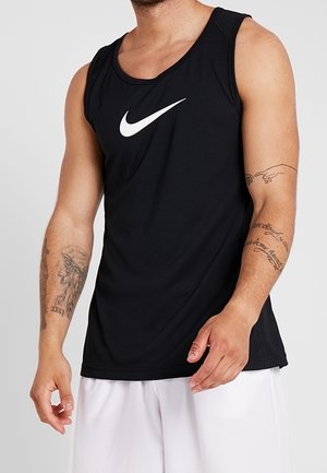 Mężczyzna w czarnym bezrękawniku Nike i białych szortach z widocznymi tatuażami na obu przedramionach na neutralnym tle.