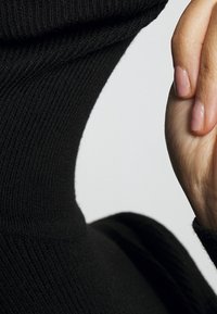 Zwarte geribbelde gebreide stof van dichtbij met een hoge kraag en een hand die de halslijn vasthoudt. Smooth textuur met subtiele variaties in het garen.