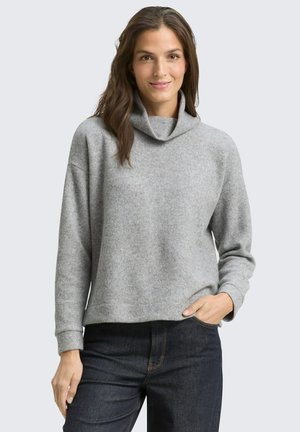 Frau trägt einen hellgrauen Rollkragenpullover mit langen Ärmeln und dunkelblaue Jeans, eine Hand in der Tasche, lächelt in die Kamera.