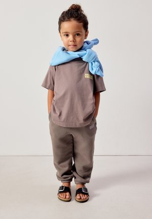 Laps seisab käed taskutes, tal on seljas pruun T-särk, sobivad püksid, mustad sandaalid ja hele sinine dressipluus õlgadel.