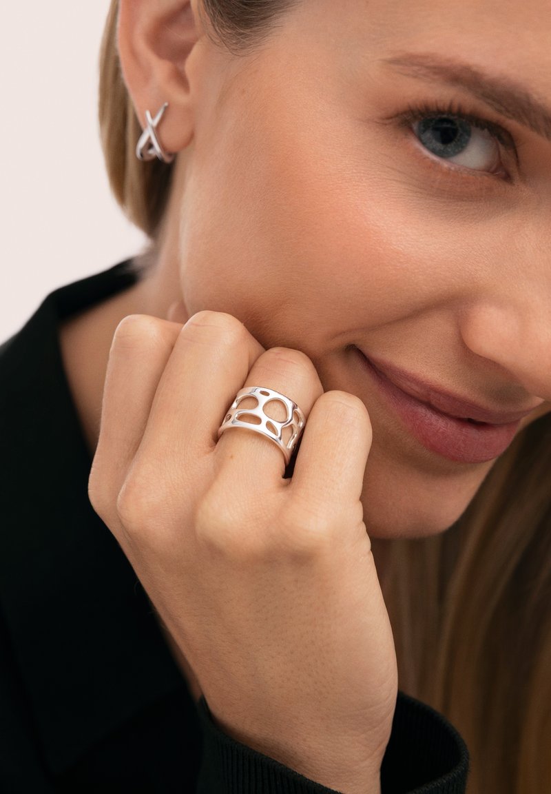 Anillo de plata con diseño recortado y orgánico, llevado en un dedo; acompañado de un pendiente simple de plata, ambos contra un fondo neutro.