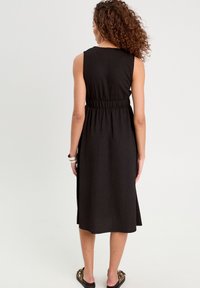 Robe sans manches noire avec une taille froncée, longueur genou, tissu texturé et fentes latérales. Portée avec des sandales plates à imprimé animal.