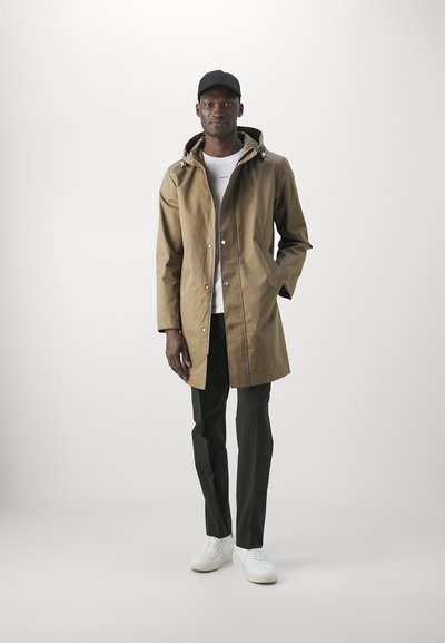 Impermeabile khaki a lunghezza ginocchio con cappuccio regolabile, bottoni a scatto e tasche laterali. Indossato sopra una camicia bianca e pantaloni neri, abbinato a sneakers bianche.