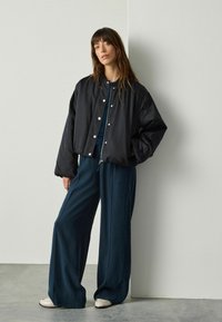 Vrouw leunend tegen een muur, gekleed in een zwarte oversized bomberjack, marineblauwe wijde broek en lichtbeige schoenen op een grijze vloer.