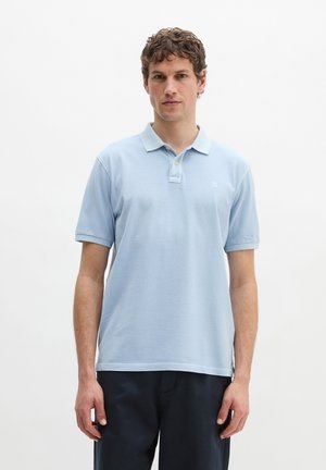 Man staat rechtop en draagt een lichtblauw poloshirt met korte mouwen en een donkere broek tegen een effen witte achtergrond.