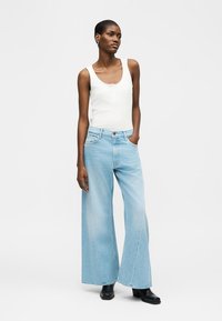 Naine, kellel on valge varrukateta särk, helerohelised laiapõlvelised teksad ja mustad pahkluu saapad, seisab ühe käega taskus.