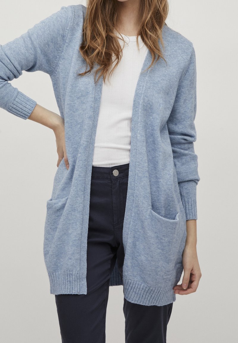 Femme portant un cardigan bleu clair ouvert avec des poches, un haut blanc côtelé et un pantalon foncé sur un fond uni.