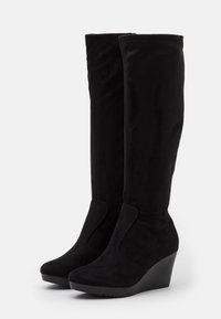 Bottes montantes noires à talon compensé, en daim doux, présentant un design épuré et une texture lisse avec des panneaux latéraux élastiques pour un enfilage facile.