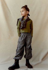 Un copil îmbrăcat cu un top verde măsliniu cu mânecă lungă, pantaloni cargo gri cu buzunare și cizme negre până la gleznă; o eșarfă gri aruncată pe gât.