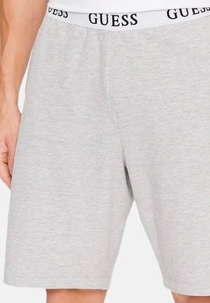 Homme portant un short gris clair avec une ceinture élastique blanche avec le logo noir "GUESS" répété.