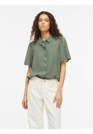 Femme aux cheveux bouclés portant une chemise à manches courtes bleu vert doux et un pantalon blanc taille haute, debout devant un fond blanc.