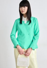 Turquoise blazer met golvende randen en dubbele knoopsluiting, gecombineerd met glanzende gele broeken en een witte parelhandtas.