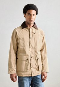 Beige jakke med krage av brunt kordfløyel, trykknapper og to frontlommer. Mykt, teksturert stoff; avslappet passform; uformelt design.