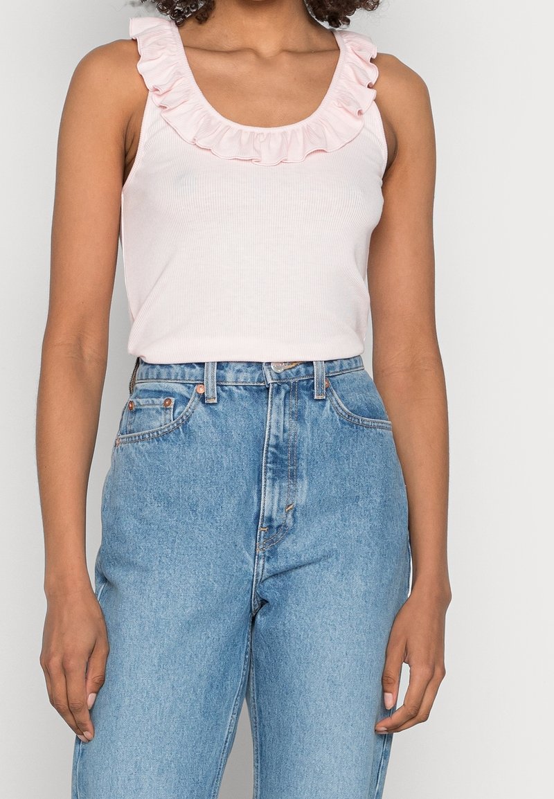 Débardeur rose clair à côtes avec un col à volants, associé à un jean en denim bleu taille haute avec une texture décolorée et une fermeture à boutons.