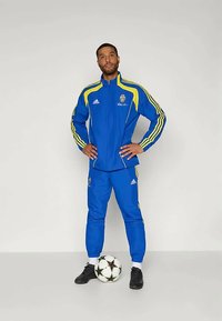 Niebieski dres z żółtymi akcentami i paskami, z bluzą na zamek i spodniami z elastycznym pasem, w połączeniu z czarnymi butami piłkarskimi i piłką.