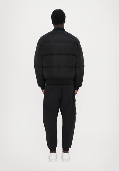 Zwarte pufferjack met horizontaal gestikte secties, ribbelboorden en onderkant. Combinatie met zwarte cargobroek en witte sneakers.