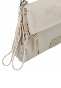 Beige Canvas-Tasche mit einem umklappbaren Deckel, strukturiertem Oberflächenmaterial, verstellbarem Riemen, Reißverschluss und dekorativen Quasten.