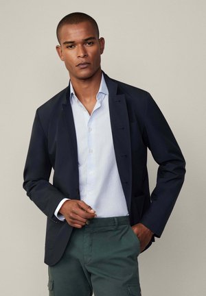 Blazer marino con cierre de un solo botón sobre una camisa de botones de color azul claro. Presenta pantalones cargo verdes que muestran un ajuste relajado y bolsillos.