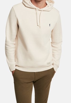 Mann trägt einen cremefarbenen Hoodie mit kleinem dunklem Logo auf der Brust und olivgrünen Hosen, die Hände in den Taschen.