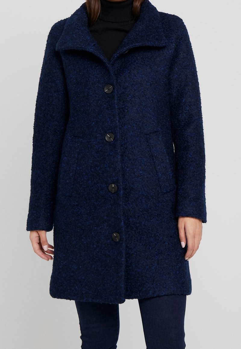 Manteau en laine bleu marine avec une finition texturée, grand col, cinq boutons noirs et poches latérales. Manches longues et coupe longueur genou.