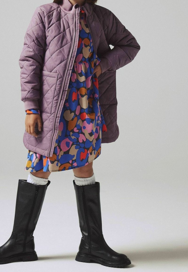 Veste matelassée lavande, robe colorée à motifs de cœurs, bottes noires montantes jusqu'aux genoux, et chaussettes blanches jusqu'aux genoux. La tenue associe textures et couleurs.
