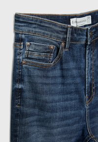 Gros plan sur un jean en denim bleu montrant une poche avant, une boucle de ceinture, des rivets métalliques, et une étiquette de taille marquée 38 de la marque Stradivarius.
