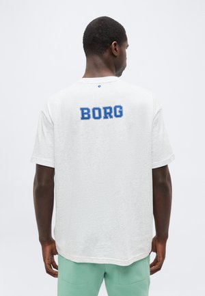 Άνδρας που φοράει λευκό κοντομάνικο πουκάμισο με μπλε κείμενο "BORG" στο πάνω μέρος της πλάτης, σε συνδυασμό με ανοιχτό πράσινο παντελόνι, γυρισμένος με την πλάτη.