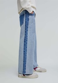 Jeans de pierna ancha de color azul claro con paneles laterales en azul oscuro y detalle en el bolsillo trasero, combinados con zapatillas grises y blancas.