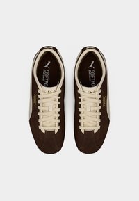 Ζευγάρι καφέ σουέτ αθλητικά παπούτσια Puma με μπεζ κορδόνια και λογότυπο Puma, εμφανιζόμενα από πάνω σε λευκό φόντο.