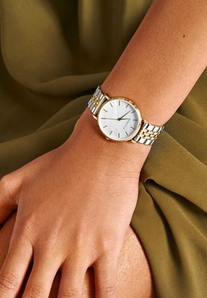 Montre-bracelet en métal argenté et doré avec un cadran blanc, des marquages minimalistes et une finition polie, portée sur une main contre un tissu olive.
