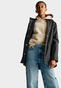 Sort vandtæt jakke med trykknapper, båret over en beige hoodie og blå denimjeans. Stoffet ser glat ud med en afslappet pasform.