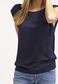 Top senza maniche blu navy con una texture liscia, scollo rotondo e dettagli arricciati sulle spalle, abbinato a jeans azzurri.